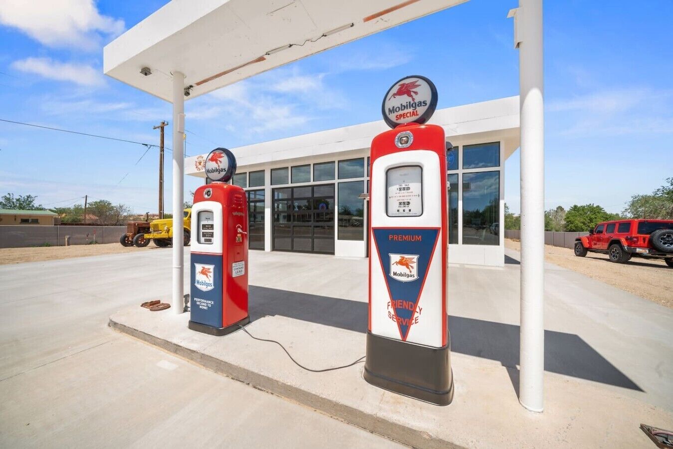 A Mobilgas gas station with two pumps, a building in the background, and a red vehicle parked nearby.