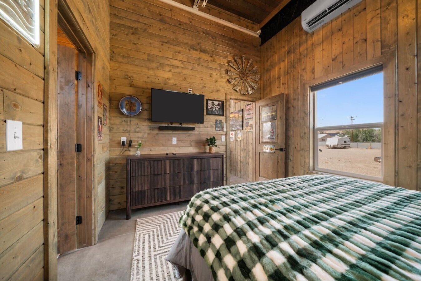 A wooden cabin bedroom with a bed, dresser, TV, and a large window.