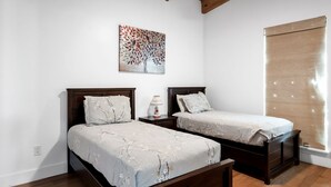 A bedroom with two beds, a wooden ceiling, a wall-mounted painting, and a window with blinds.