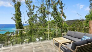 Ein Balkon mit einer Couch und einem Tisch, mit Blick auf eine Küstenlandschaft mit Bäumen und Meer.