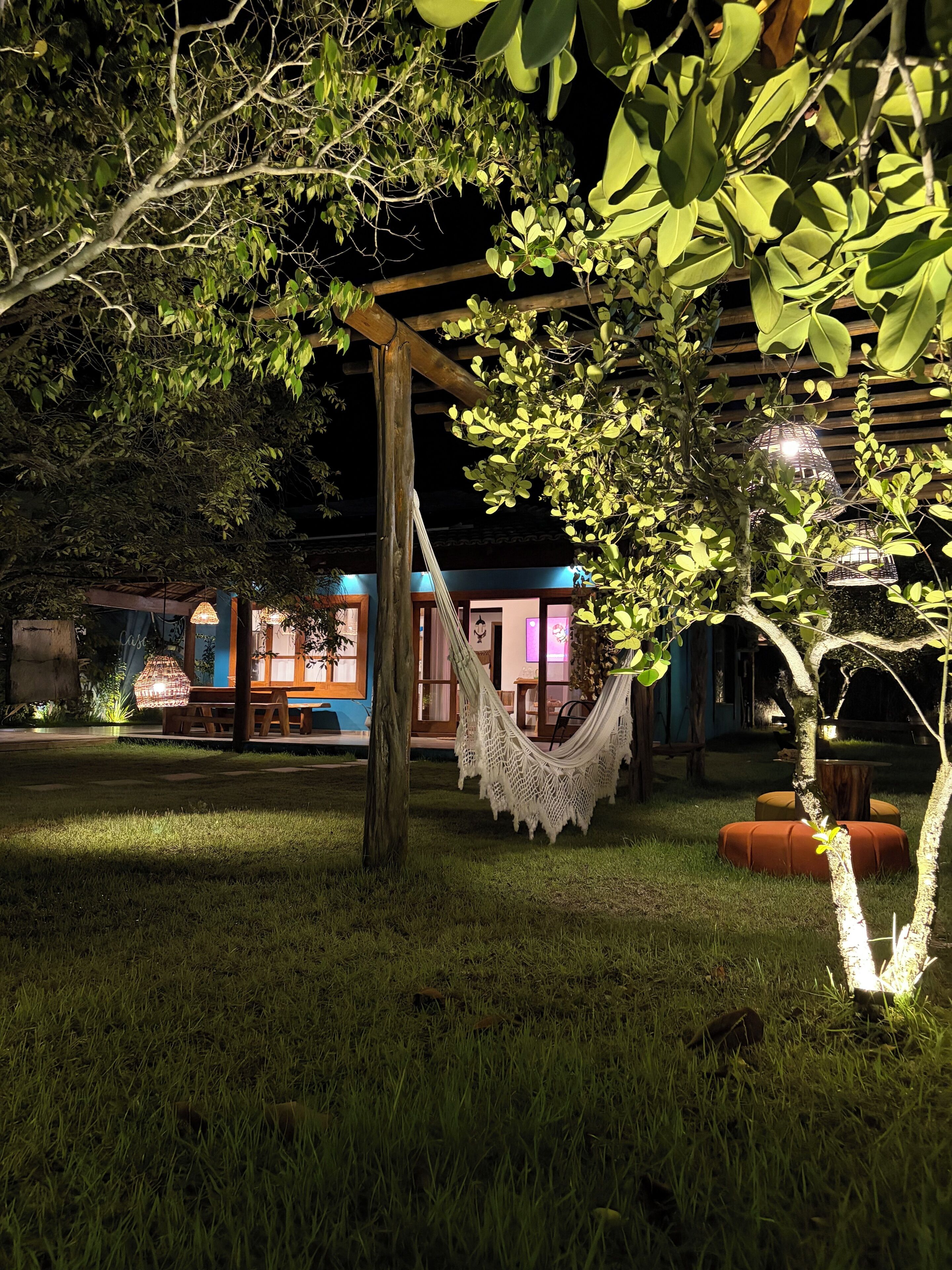 Un hamac sous un arbre, une pergola en bois et, au fond, un bâtiment dont les fenêtres sont illuminées.
