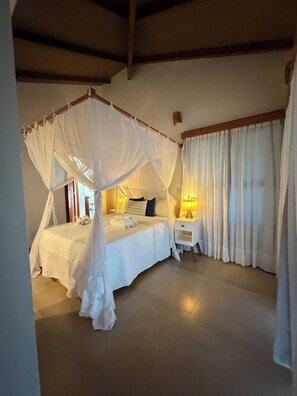 A bedroom with a four-poster bed, a bedside table with a lamp, and a window with curtains. - Humpback Whale House Azul (Caraíva)