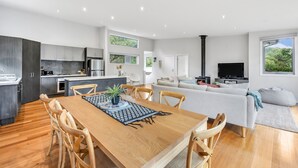 A modern kitchen with dark cabinets, a dining area with a wooden table and chairs, a living room with a sofa and a TV, and a large window with a view of greenery. - Fairhaven Treetops Lookout (Fairhaven)