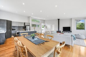 Eine moderne Küche mit dunklen Schränken, ein Essbereich mit Holztisch und Stühlen, ein Wohnzimmer mit einer Couch und einem Fernseher sowie ein großes Fenster mit Blick auf Grünflächen.