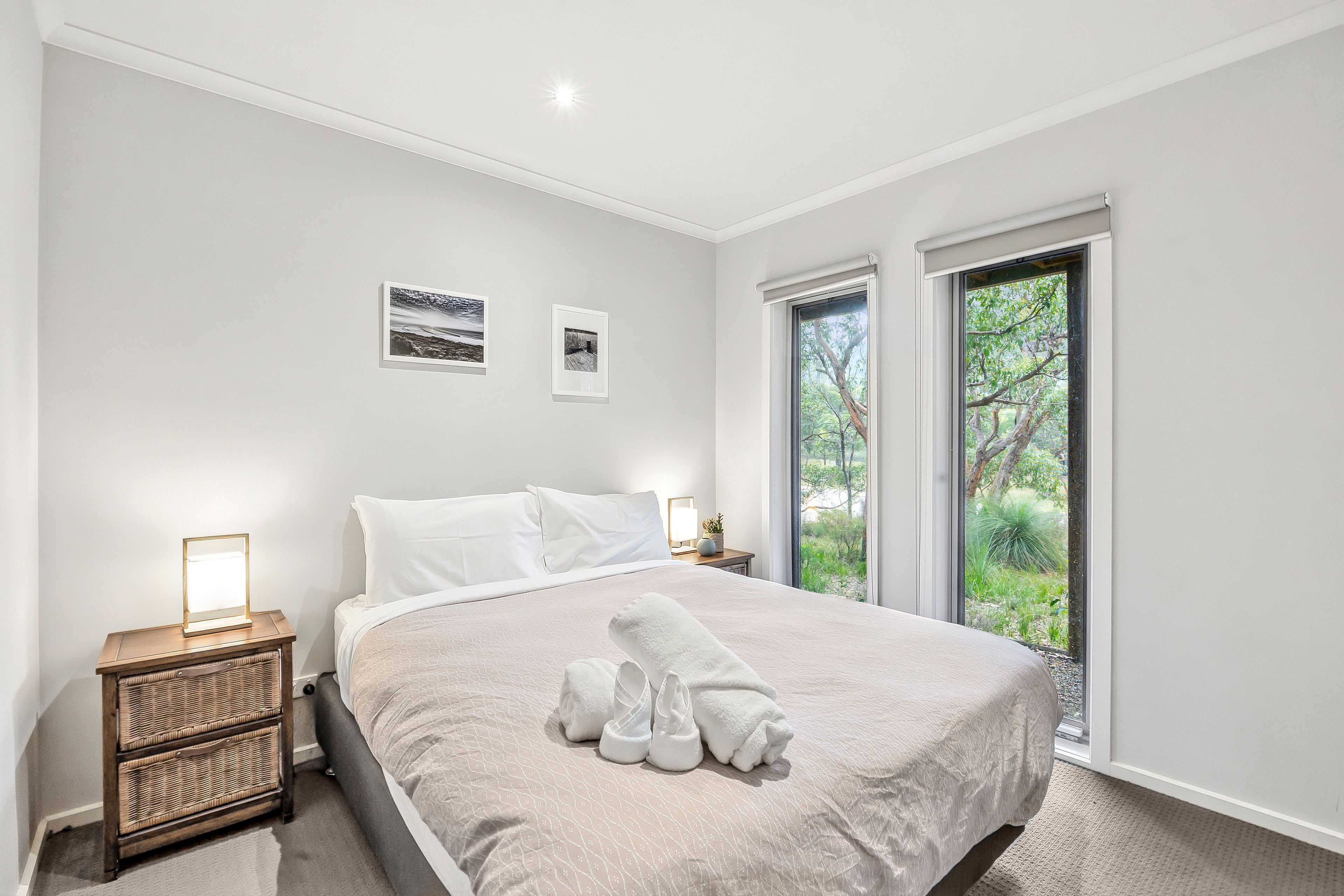 Ein Schlafzimmer mit einem Bett, zwei Fenstern, einem Nachttisch und zwei gerahmten Bildern an der Wand.