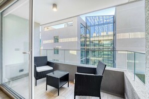 A balcony with wicker furniture, a glass railing, and a view of a modern building. - Estoril Suite | Clínica y Centro Comercial (Las Condes)