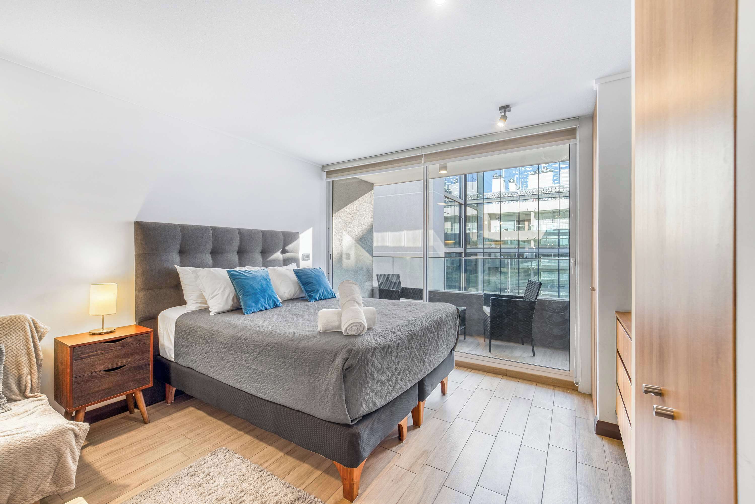 Een moderne slaapkamer met een groot bed, een nachtkastje, een stoel en uitzicht op de stad door een groot raam.