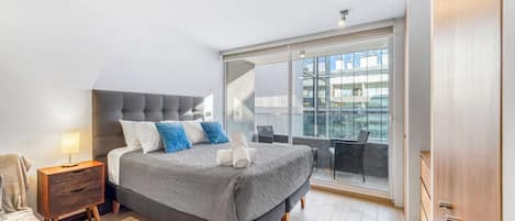 A modern bedroom with a large bed, a nightstand, a chair, and a view of the city through a large window.