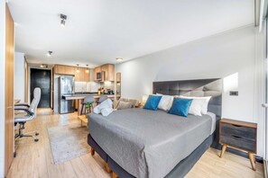A modern bedroom with a large bed, a desk, and a kitchenette. - Estoril Suite | Clínica y Centro Comercial (Las Condes)