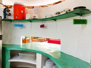 A kitchen with a green countertop, a red water dispenser, and a white blender. There are hanging labels for 'ALEGRIA' and 'NUTRITION'.