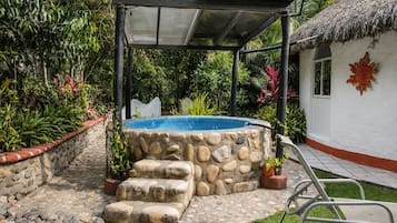 Tinaja de piedra con una pérgola de metal, rodeada por un jardín con diversas plantas y una estructura de techo de paja.