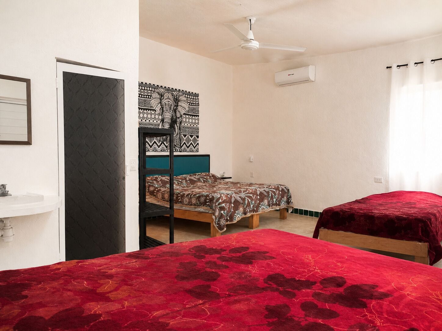 A room with a bunk bed, a single bed, a ceiling fan, an air conditioning unit, and a window with curtains.