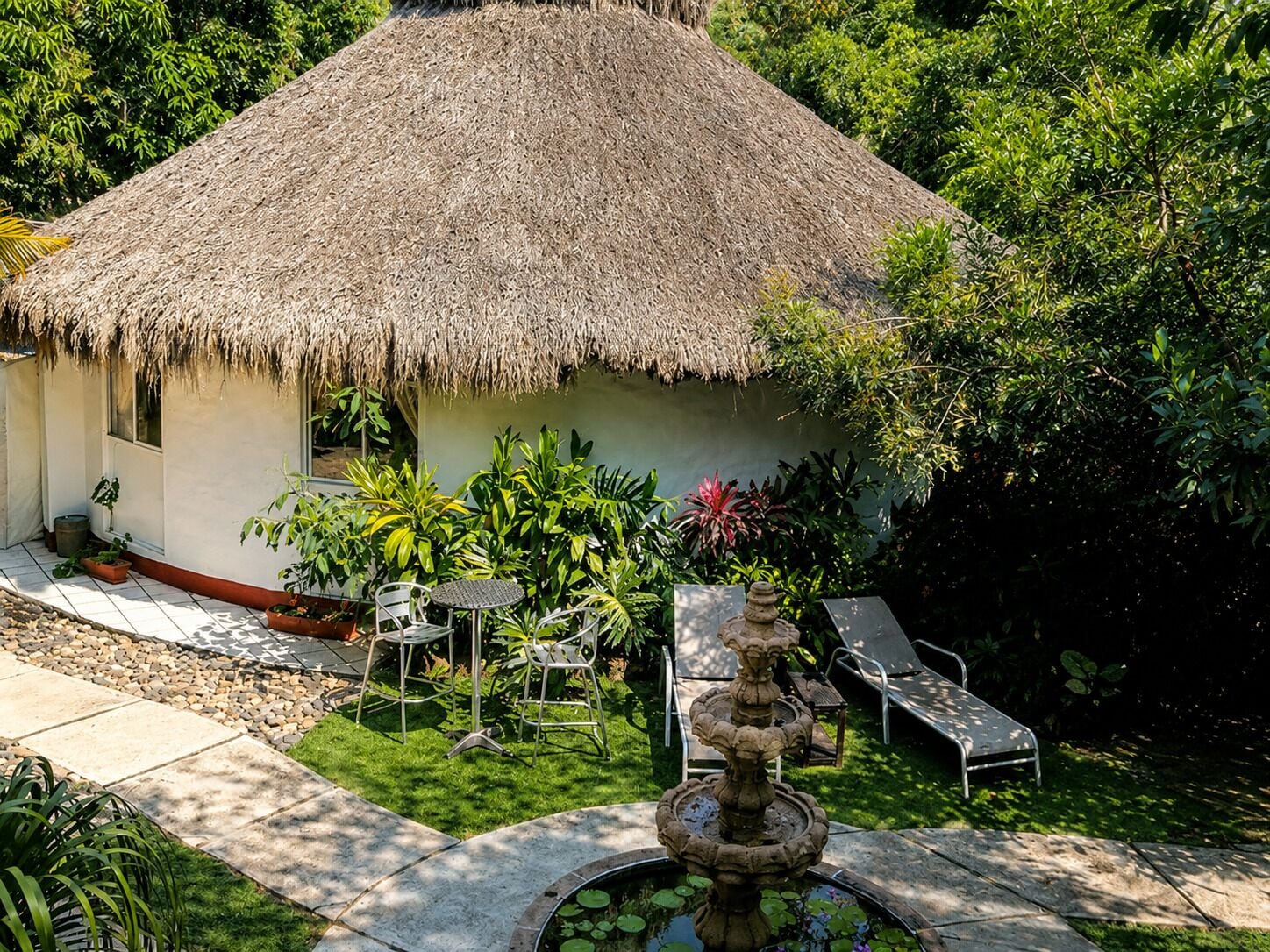 Una casa con techo de paja, un jardín, asientos exteriores y una fuente.