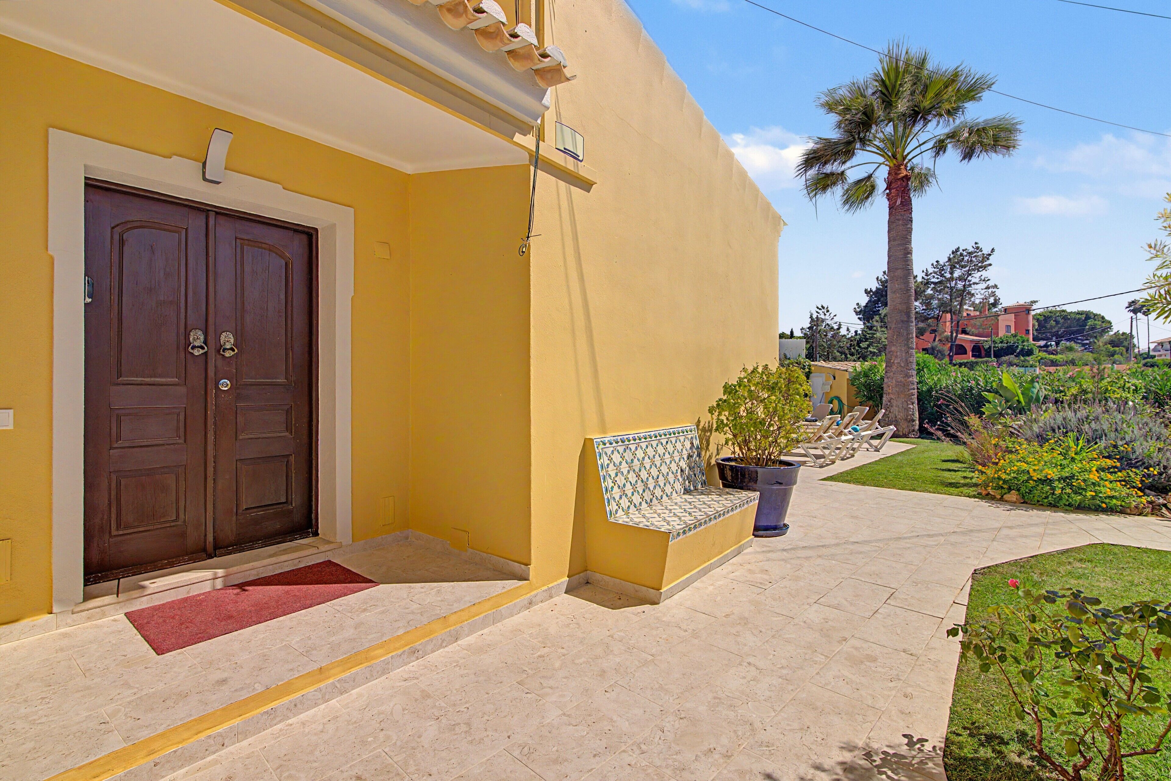 Un edificio amarillo con puerta marrón, una banca y una palmera.