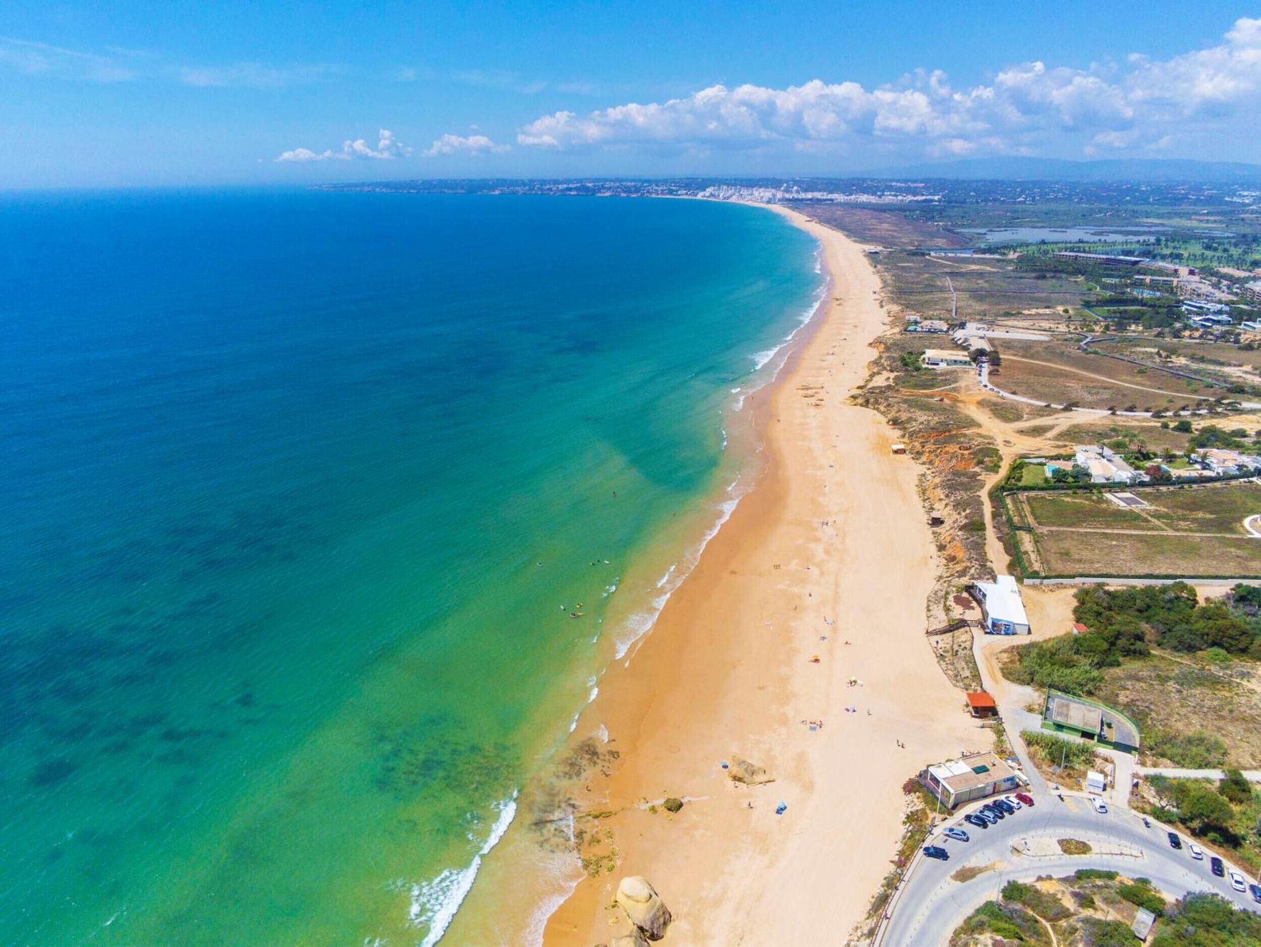 Vue aérienne d’une zone côtière avec une plage, des eaux d’un bleu limpide et une route bordée de voitures garées.