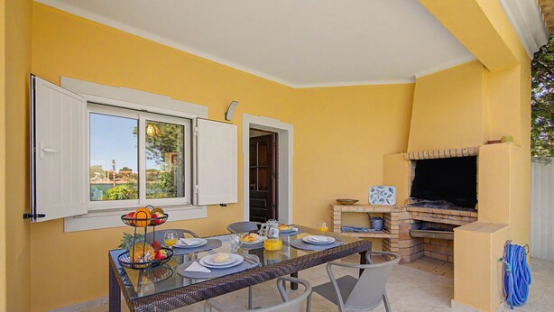 Un coin repas avec une table dressée pour quatre personnes, un barbecue et une fenêtre avec des rideaux.