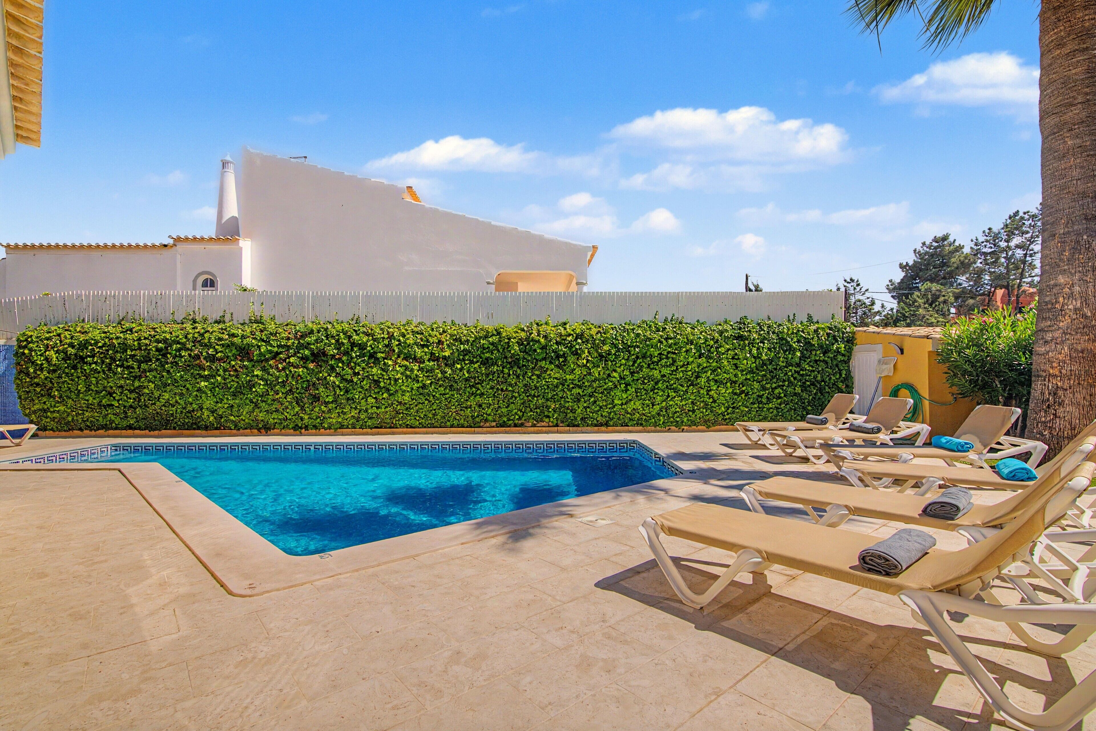 Piscina com espreguiçadeiras, um edifício branco ao fundo e um conjunto de sebe.