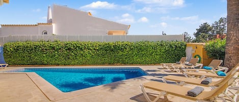 Piscina com espreguiçadeiras, um edifício branco ao fundo e um conjunto de sebe.