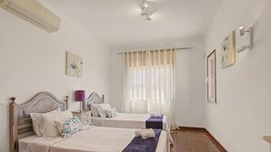 Une chambre d’hôtel avec deux lits, une fenêtre avec des rideaux légers, un climatiseur fixé au mur et une petite table avec une lampe.