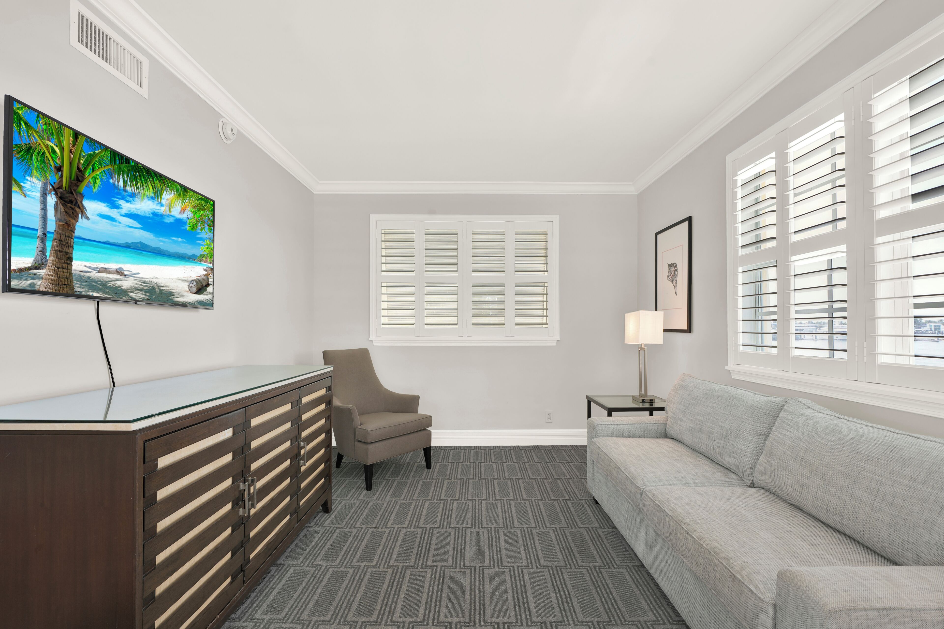 Hotelkamer met een bank, bureau met lades, stoel, raam met shutters en een schilderij van een strand, plus een lamp.
