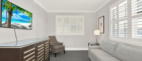 A hotel room with a sofa, a desk with drawers, a chair, a window with shutters, a painting of a beach, and a lamp.