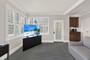 Una sala de estar con un televisor de pantalla plana sobre un mueble de entretenimiento negro, un sofá gris y una cocina con armarios de madera.