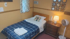 Un lit simple avec une couvre-lit à carreaux bleus et blancs, une tête de lit en bois, une table de chevet avec une lampe, un ventilateur et un mur orné de tableaux encadrés.