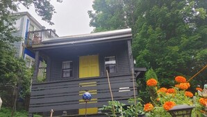 Une maison en bois à deux étages, avec une porte et des fenêtres jaunes, entourée de verdure et de fleurs orange.