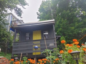Una casa de fusta de dues plantes amb una porta i finestres grogues, envoltada de verdura i flors taronja.
