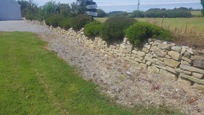 Ländliche Landschaft mit einer Steinmauer, einem Schotterweg und einem Gebäude mit einem Schild, das Hinweise nach Dingle und Tralee gibt.