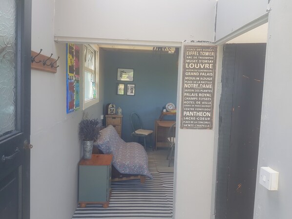 A narrow hallway with a striped floor leading to a room with a bed, a chair, and a small table. A sign listing various Parisian landmarks is visible on the wall.