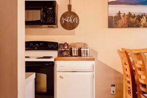 Een compacte keuken met een magnetron, fornuis en een kleine koelkast. Er is een houten bordje met de tekst 'gather' en een landschapsschilderij aan de muur.