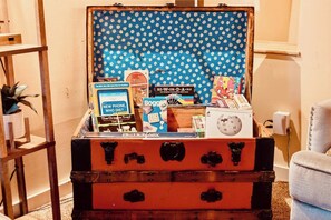 Ein alter Koffer, gefüllt mit Gesellschaftsspielen und Büchern, steht auf einem Holzboden vor einem blauen, gemusterten Hintergrund.