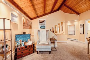 Un loft espacioso con techo alto, un área de estar central con una silla y una mesita, una escalera que lleva a un nivel superior y un estante en la pared con varios objetos.