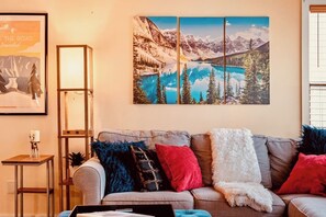 Een woonkamer met een grijze bank, kleurrijke sierkussens, een lijstwerk-landschapschilderij en een tafeltje met een lamp.
