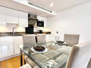 A modern kitchen with white cabinets, a black microwave, and a glass dining table with chairs. - Luxury 4 Bedrooms in Jerome House London (London)