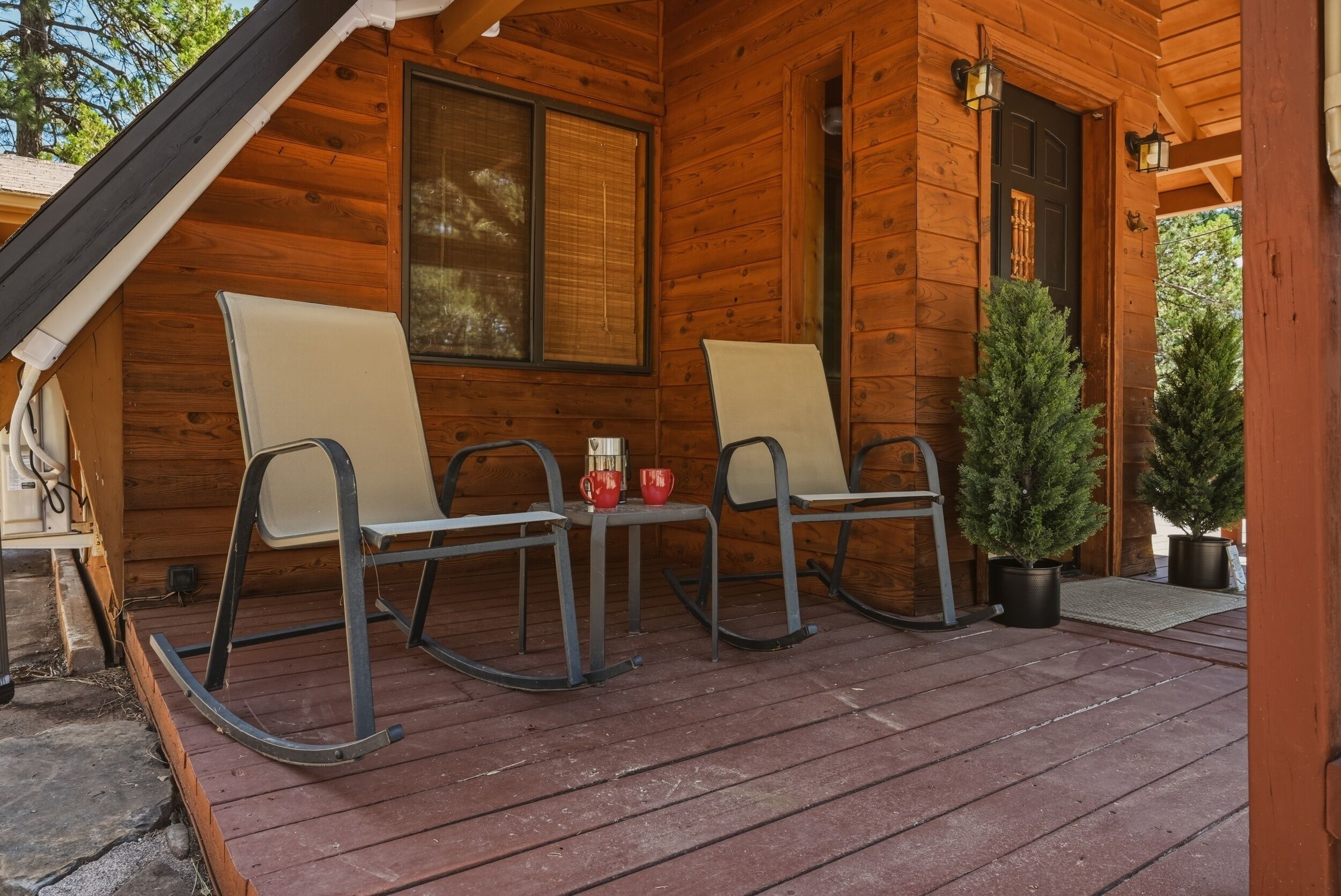 A wooden cabin with a porch, two rocking chairs, a small table with cups, and a potted tree.