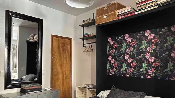 A bedroom with a bed, a wardrobe, a mirror, a shelf with books, and a floral wall. - Charming Shared Unit with Host. Beautiful Park Slope on 5th Ave (Brooklyn)