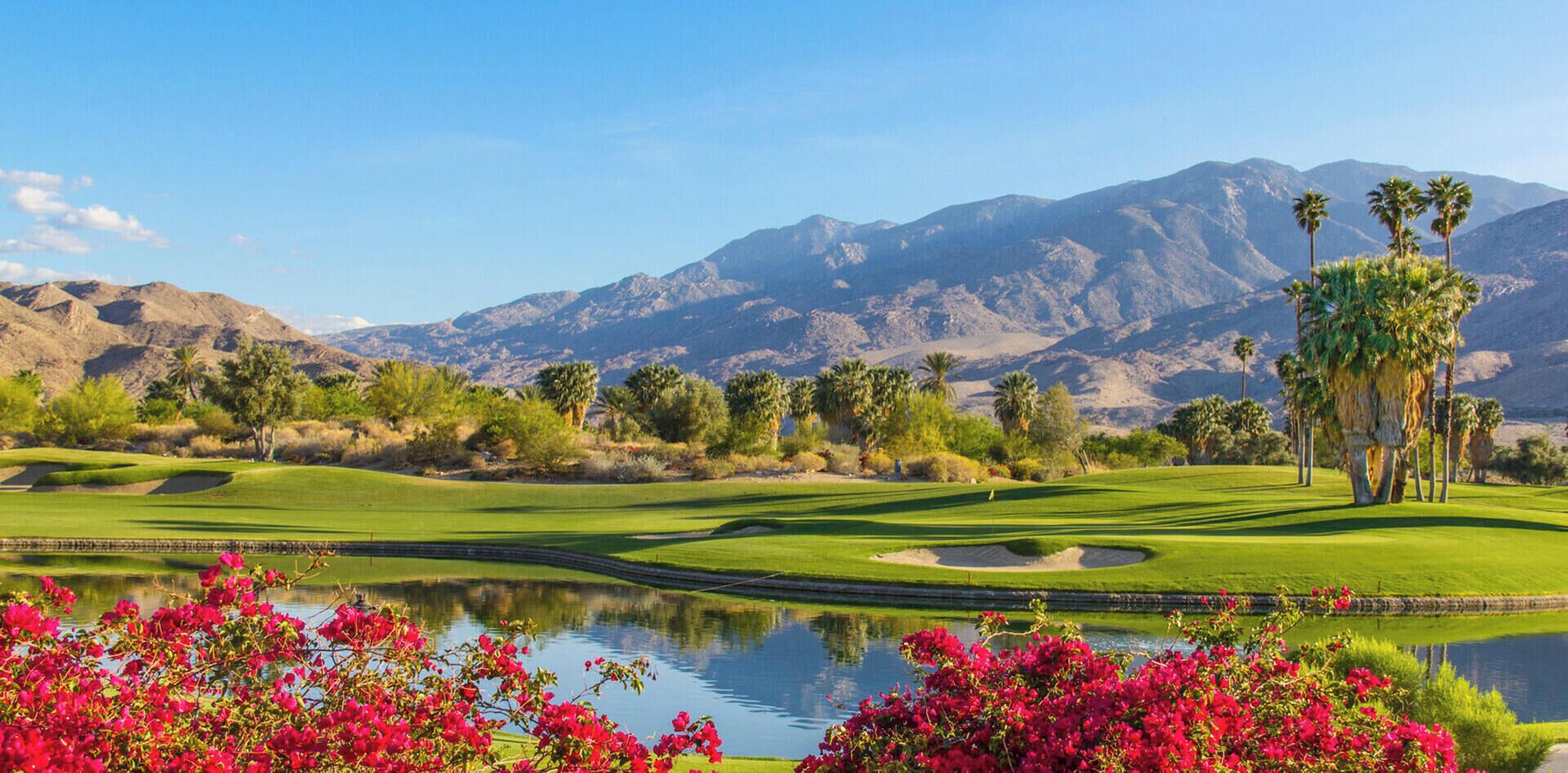 Un parcours de golf avec un étang, des palmiers et des montagnes en arrière-plan.