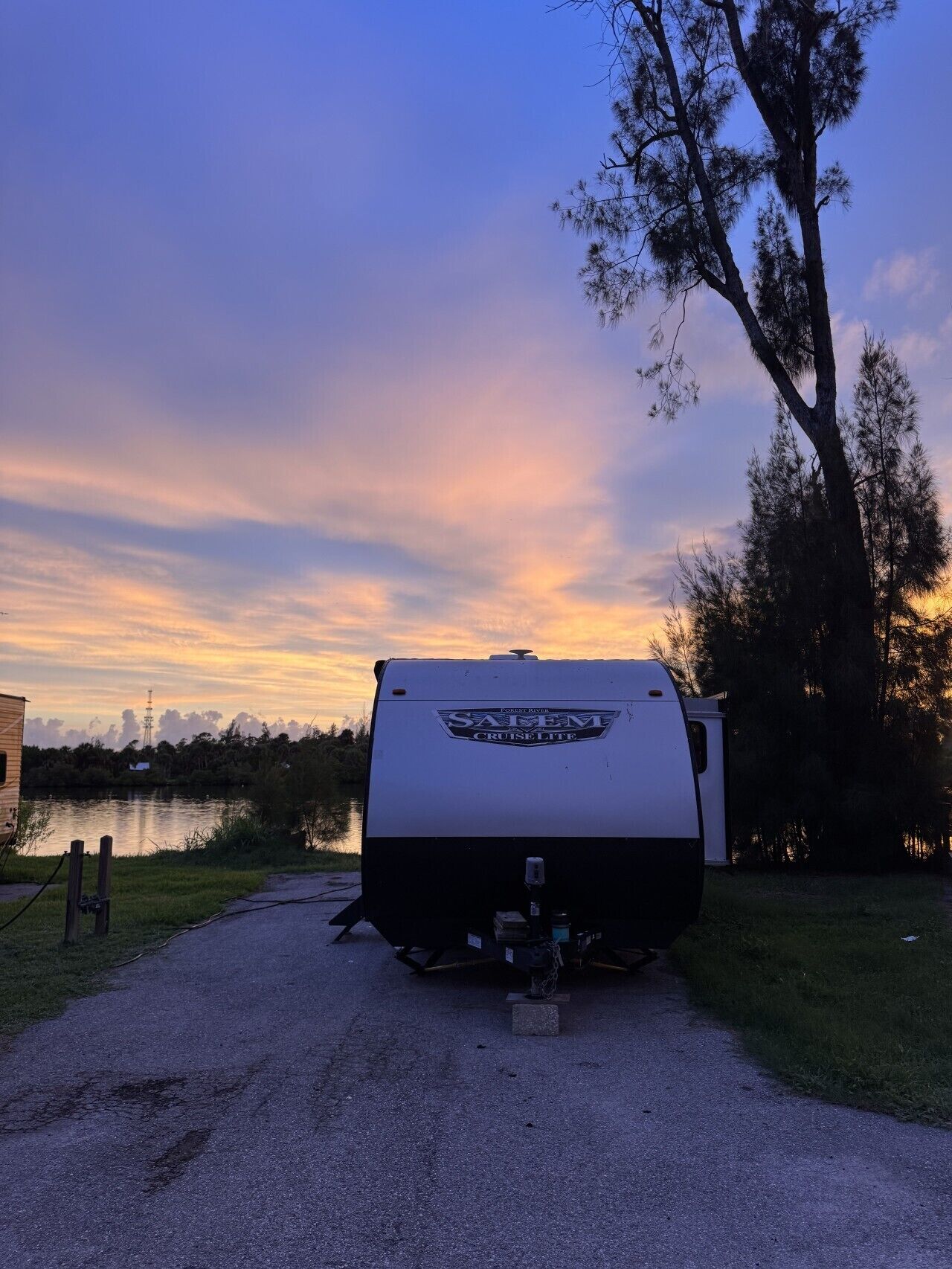 Un camping-car blanc, arborant la mention « SAVEM CRUISE LITE », est garé sur un chemin de gravier, au bord d’une étendue d’eau. On aperçoit des arbres et un bâtiment en arrière-plan.