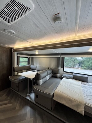 A modern RV interior with a sectional sofa, a dining table, and a window providing a view of the outdoors.