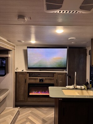 A modern RV interior with a flat-screen TV mounted above a fireplace, wooden cabinetry, and a kitchenette area.