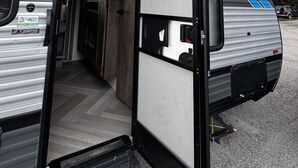 Une caravane de voyage avec une porte coulissante ouverte, révélant un intérieur doté d’un plancher en bois et d’un petit coin cuisine.