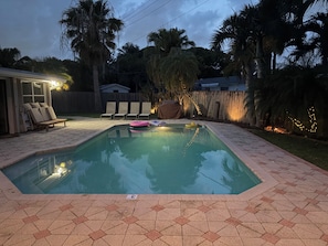 A swimming pool with lounge chairs and a covered seating area, surrounded by a wooden fence and palm trees. - The Oasis Private Pool +Gazebo 5 Minutes to the Beach! (Pompano Beach)