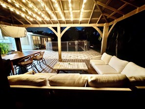 Een terras buiten met een houten tafel, stoelen, een bank en een overdekt gedeelte met lichtslingers.
