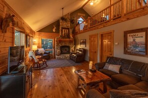 A log cabin interior with a stone fireplace, wooden furniture, and a balcony.