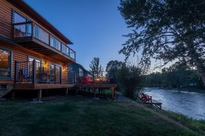 A wooden cabin with a deck overlooking a river, featuring outdoor seating and a large window.