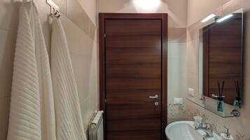Un baño con puerta de madera, un lavabo blanco, estante de vidrio con artículos de aseo y dos toallas colgadas en un perchero.