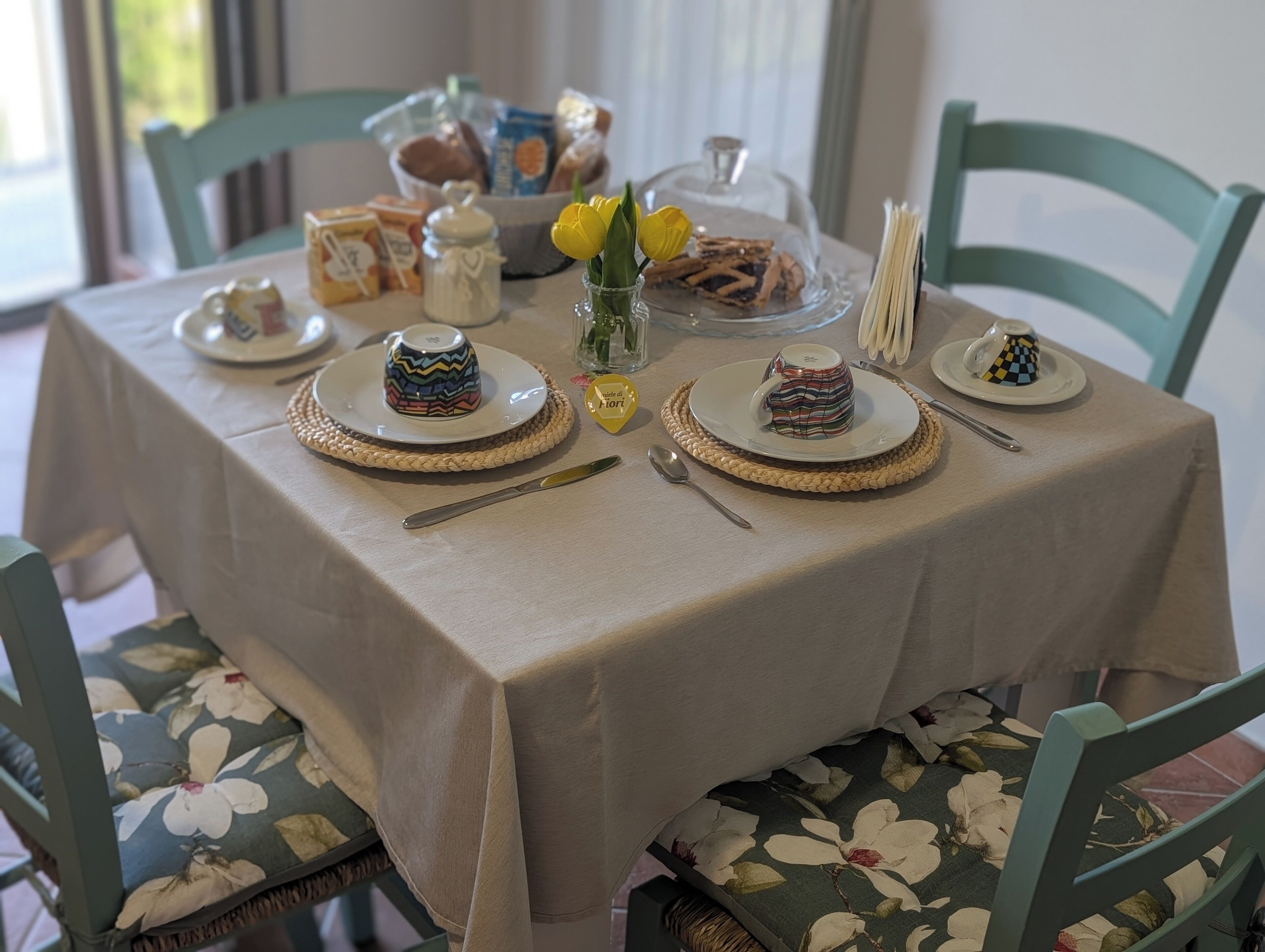 A dining table with a beige tablecloth, floral chairs, and various dishes and utensils.