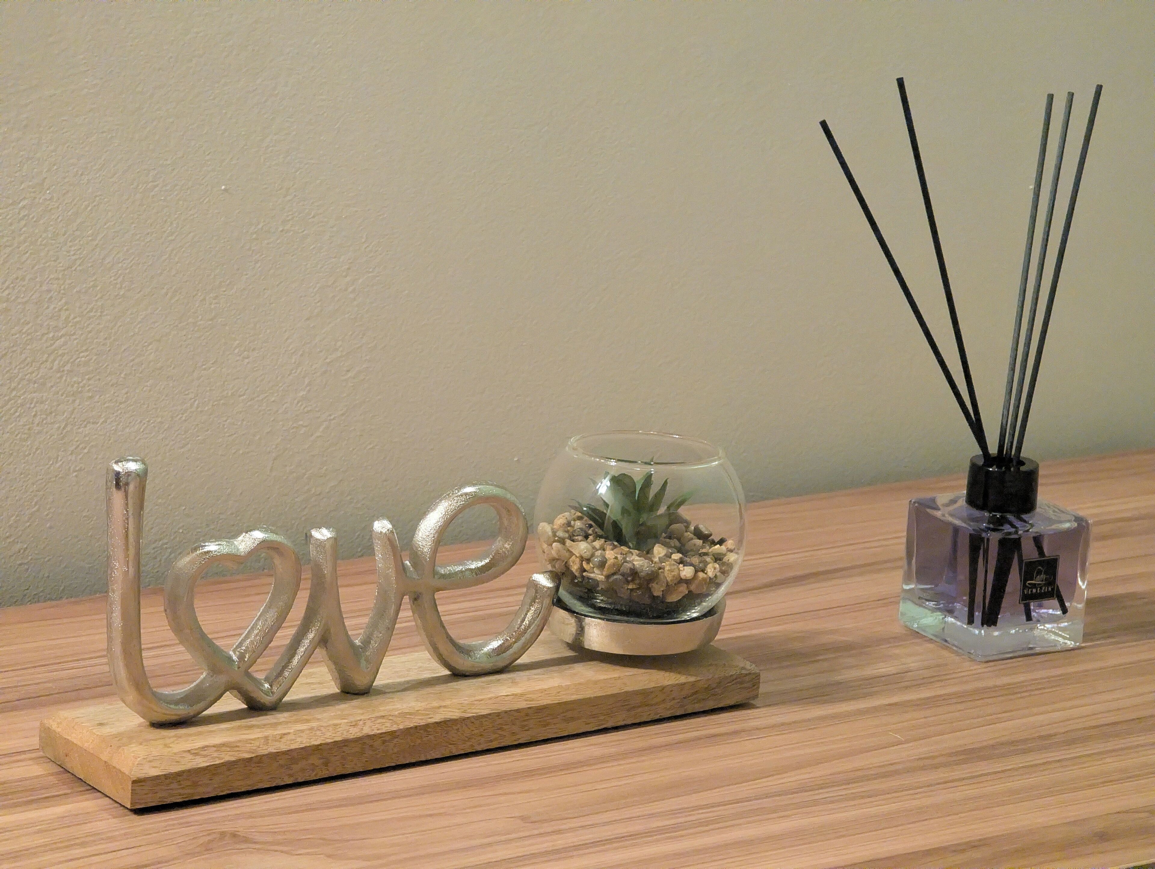 A wooden table with a decorative metal sign spelling 'love,' a small glass bowl with pebbles and a plant, and a reed diffuser.